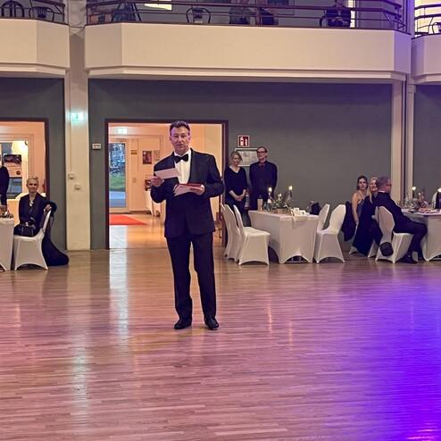 Mann im Smoking hält ein Papiermikrofon in einem festlich dekorierten Saal mit Tischen und Gästen im Hintergrund.