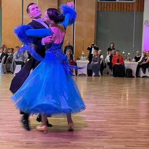 Tanzpaar in eleganter Kleidung, der Mann führt, während die Frau mit fließendem, blauem Kleid auf einer Tanzfläche tanzt.