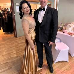 Paar in eleganter Abendkleidung posiert lächelnd, Hintergrund mit anderen festlich gekleideten Gästen.