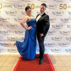 Ein Paar posiert vor einem festlichen Hintergrund mit dem Schriftzug „50 Jahre TSC Brühl“ auf einem roten Teppich.