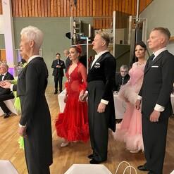 Fünf Tänzer in formeller Kleidung stehen auf einer Tanzfläche, bereit für einen Wettkampf.