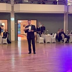 Mann im Smoking hält ein Papiermikrofon in einem festlich dekorierten Saal mit Tischen und Gästen im Hintergrund.