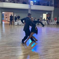 Tanzpaar in eleganter Kleidung auf einer Tanzfläche, umgeben von Zuschauern und beleuchtet von bunten Lichtern.