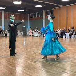 Tanzpaar in festlicher Kleidung bewegt sich auf einer Tanzfläche, umgeben von Publikum und Tischdekorationen.