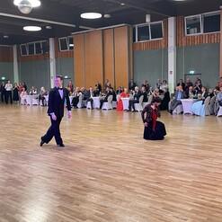 Tanzpaar in eleganter Kleidung auf einer Holzfläche in einem festlich dekorierten Saal, Publikum im Hintergrund.