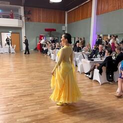 Tänzerin in gelb-goldenem Kleid auf einer Tanzfläche, Zuschauer sitzen an Rändern und beobachten die Darbietung.