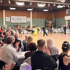 Tanzpaar in eleganter Kleidung auf einer Tanzfläche vor applaudierendem Publikum in einem festlichen Saal.