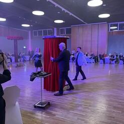 Zwei Männer und eine Frau stehen vor einem roten Vorhang auf einer Veranstaltung; Gäste sitzen an Tischen im Hintergrund.