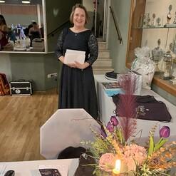 Eine lächelnde Frau in einem schwarzen Kleid steht neben einem Tisch mit einer Blumenarrangement und Trophäen im Hintergrund.