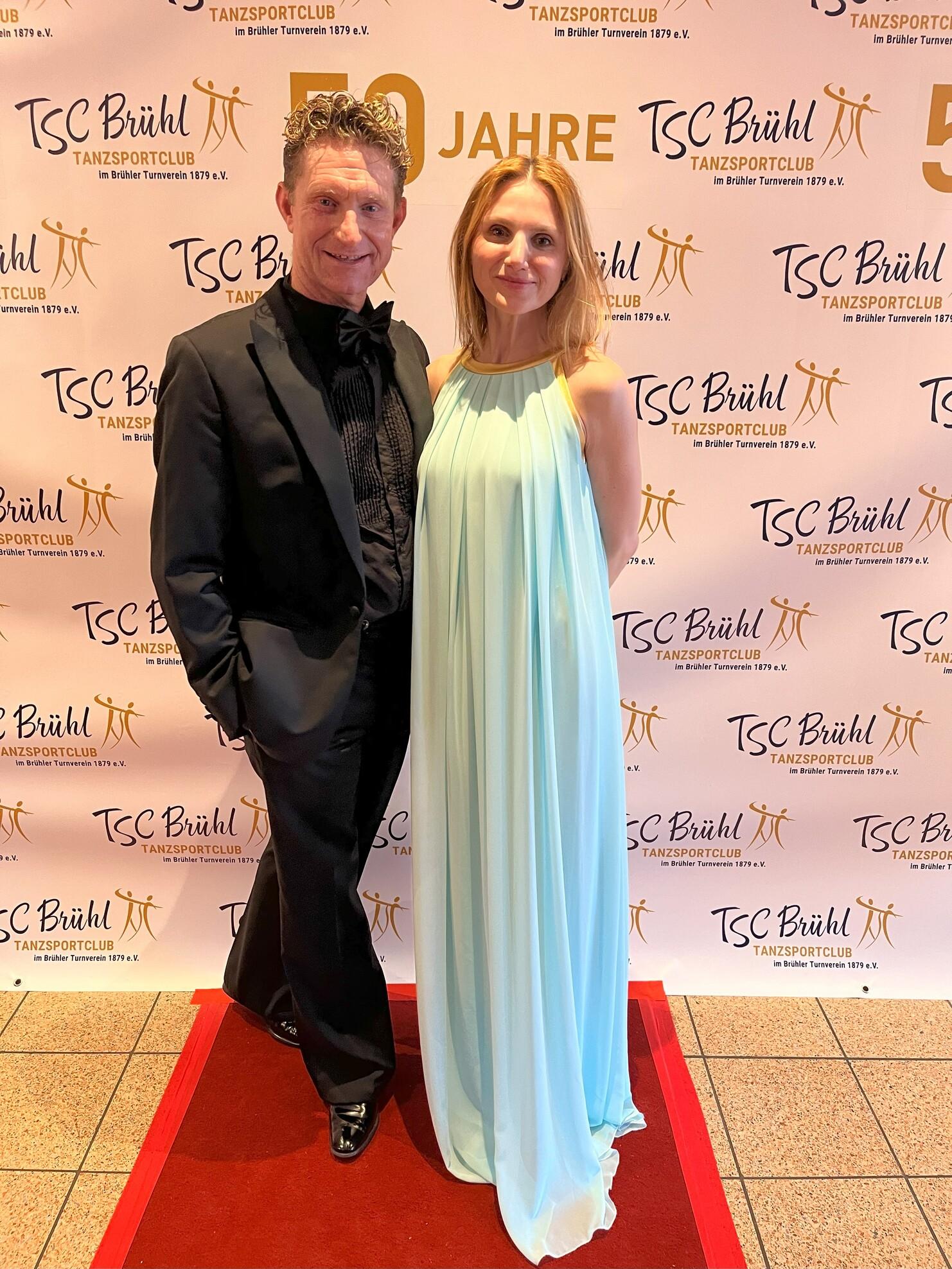 Paar in festlicher Kleidung steht vor einem Banner zum 50-jährigen Jubiläum des TSC Brühl.
