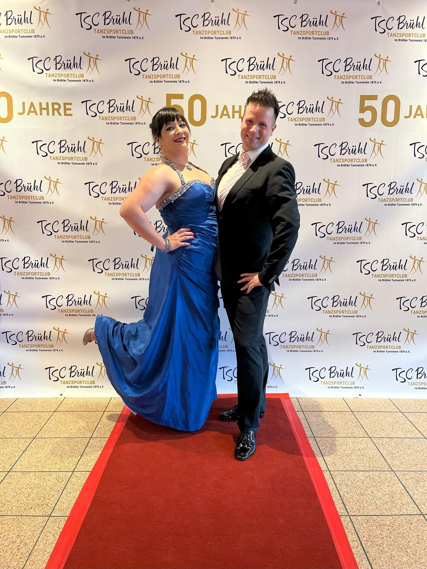 Ein Paar posiert vor einem festlichen Hintergrund mit dem Schriftzug „50 Jahre TSC Brühl“ auf einem roten Teppich.