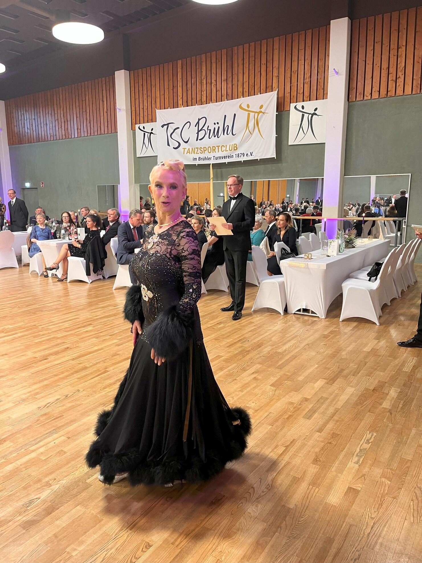 Eine Tänzerin in einem eleganten, schwarzen Kleid mit Pailletten und Pelzverzierungen steht in einem festlichen Saal.