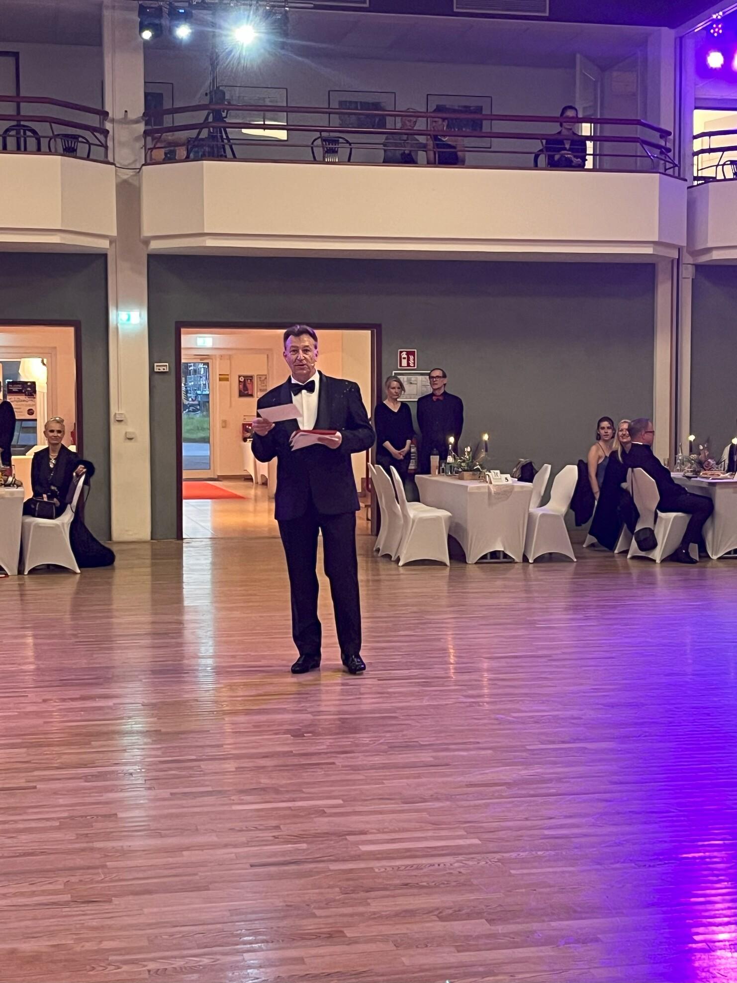 Mann im Smoking hält ein Papiermikrofon in einem festlich dekorierten Saal mit Tischen und Gästen im Hintergrund.