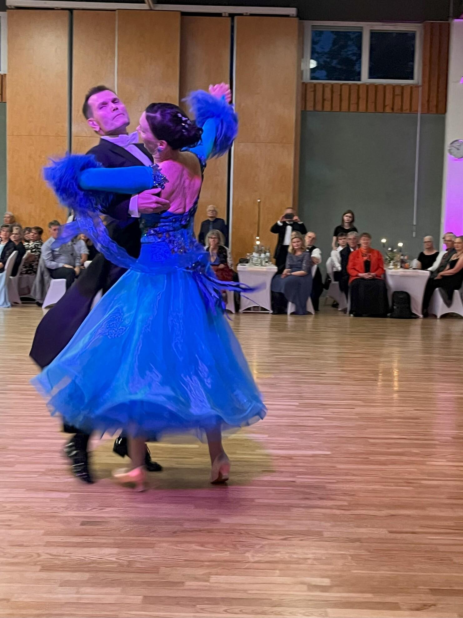 Tanzpaar in eleganter Kleidung, der Mann führt, während die Frau mit fließendem, blauem Kleid auf einer Tanzfläche tanzt.