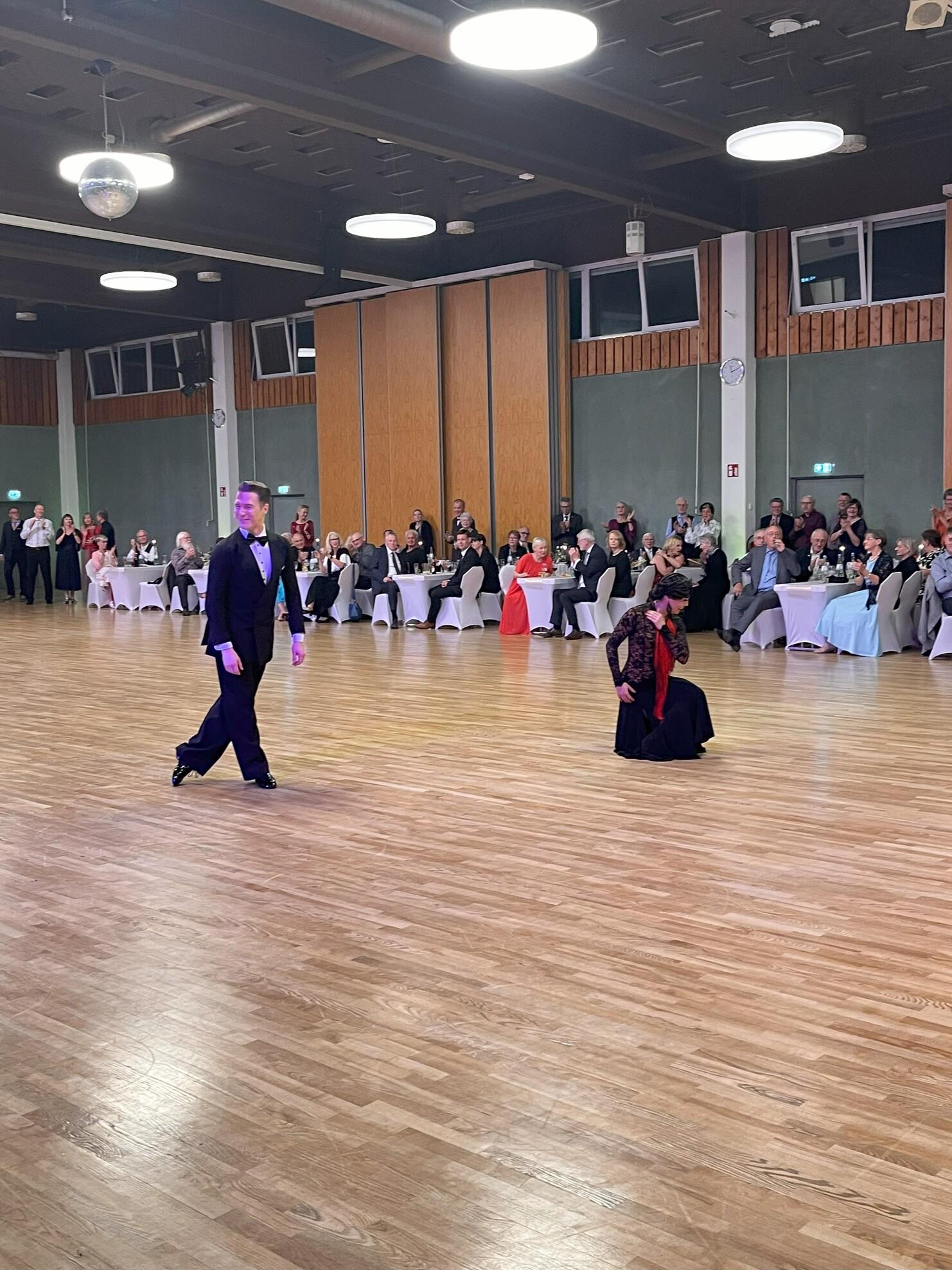 Tanzpaar in eleganter Kleidung auf einer Holzfläche in einem festlich dekorierten Saal, Publikum im Hintergrund.