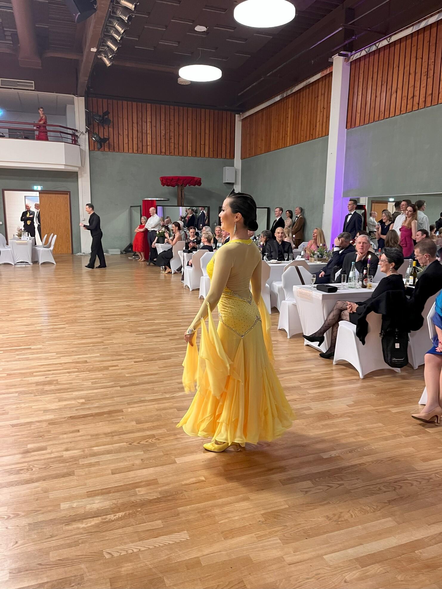 Tänzerin in gelb-goldenem Kleid auf einer Tanzfläche, Zuschauer sitzen an Rändern und beobachten die Darbietung.