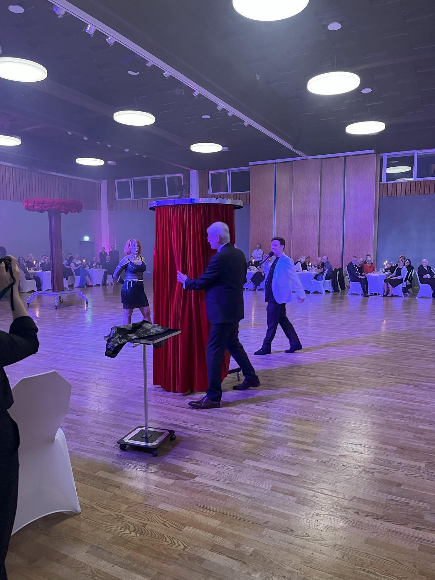 Zwei Männer und eine Frau stehen vor einem roten Vorhang auf einer Veranstaltung; Gäste sitzen an Tischen im Hintergrund.