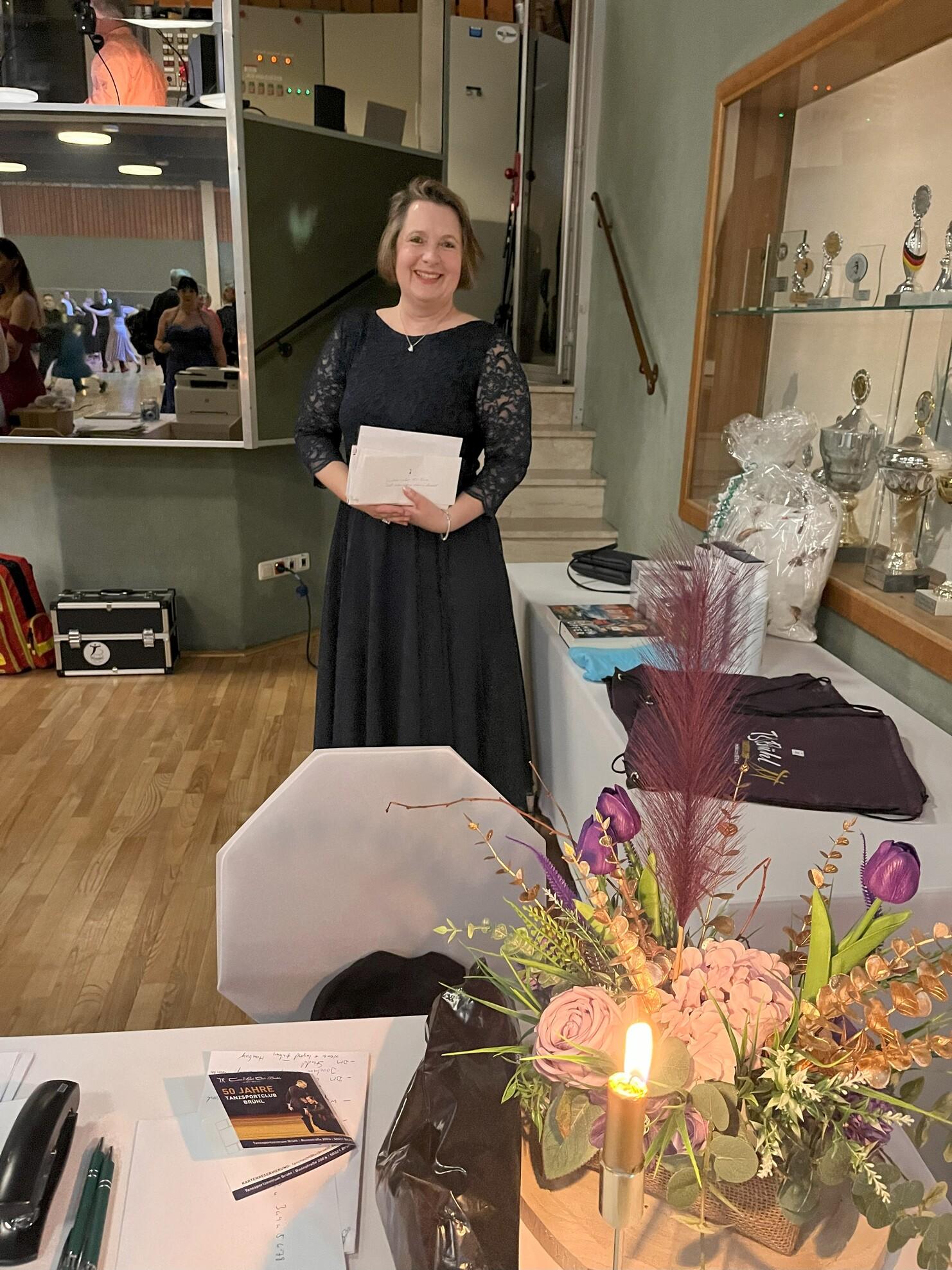 Eine lächelnde Frau in einem schwarzen Kleid steht neben einem Tisch mit einer Blumenarrangement und Trophäen im Hintergrund.