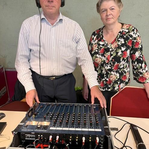 Zwei Personen stehen neben einem Audiomischpult, eins trägt Kopfhörer, beide blicken in die Kamera.