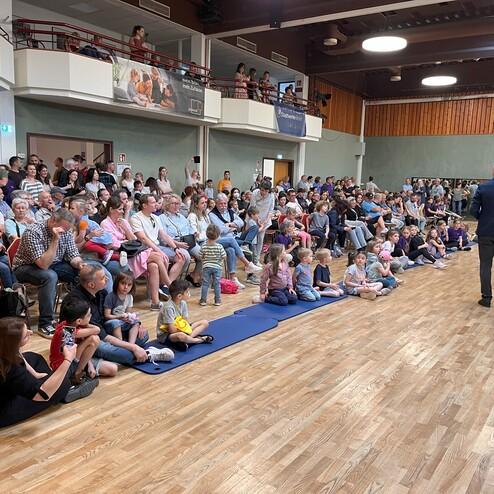 Große Menschenmenge sitzt auf Stühlen und Matten in einem Saal, während ein Redner auf der Bühne spricht.