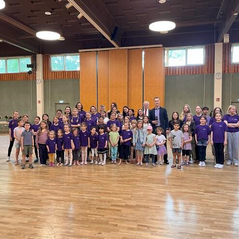 Gruppenfoto von Kindern und Erwachsenen in lila T-Shirts in einem großen Raum mit Holzfußboden.