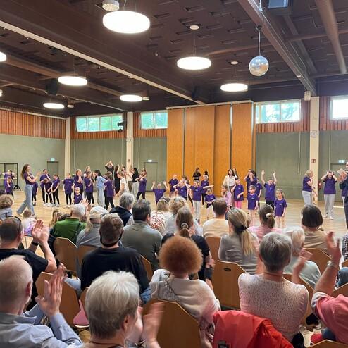 Tanzaufführung in einer Halle mit Kindern in lila Shirts, während das Publikum applaudiert.