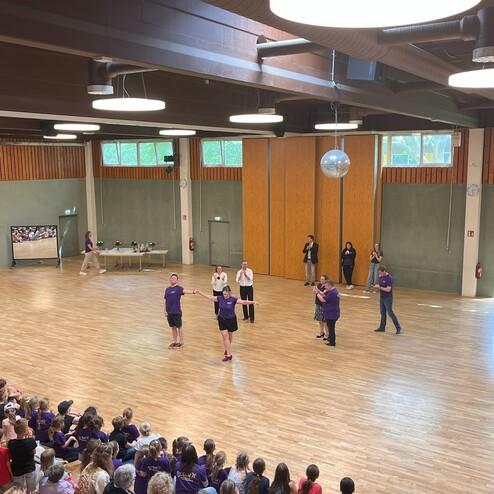 Tanzen auf einer Bühne: Teilnehmer in lila T-Shirts präsentieren eine Choreografie vor einem applaudierenden Publikum.