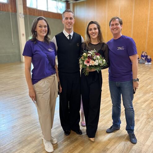 Vier Personen posieren in einem hellen Raum; zwei tragen lila T-Shirts, eine hält einen Blumenstrauß.