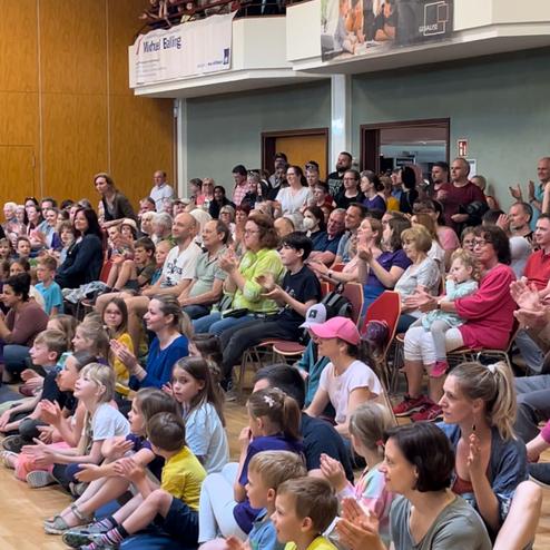 Eine große Menge von Menschen, überwiegend Kinder, sitzt auf dem Boden und applaudiert in einem Veranstaltungsraum.