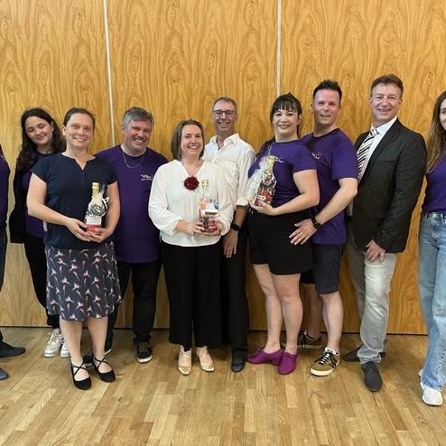 Gruppenportrait von zehn Personen in lila T-Shirts, die Geschenke in der Hand halten, vor einer Holzverkleidung.