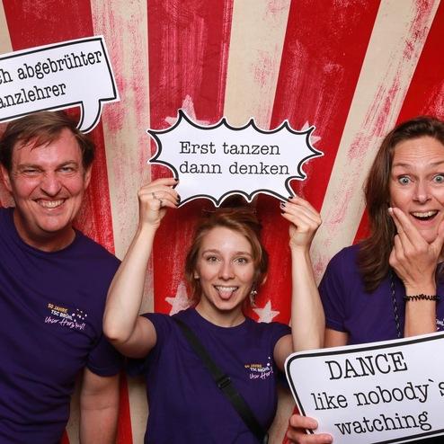 Drei Personen in lila Shirts halten Sprechblasen mit Sprüchen vor einem gestreiften Hintergrund.
