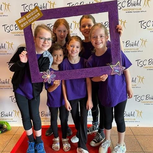 Sechs Kinder in lila T-Shirts posieren mit einem glitzernden Rahmen vor einem Banner zum 50-jährigen Jubiläum des TSC Brühl.