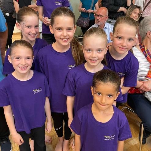 Sechs Mädchen in lila T-Shirts mit Glitzerschminke posieren lächelnd in einer Menschenmenge.