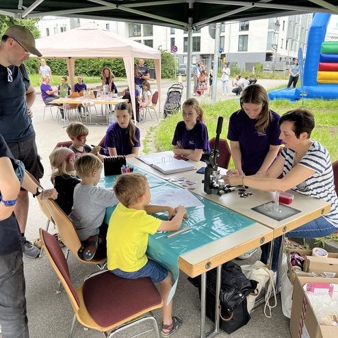 Kinder und Erwachsene am Basteltisch mit Mikroskop und Farben unter einem Zelt auf einer Au&szlig;eneventfl&auml;che.