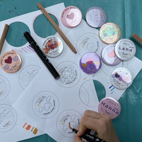 Bunt bedruckte Buttons und Zeichenskizzen liegen auf einem Tisch, w&auml;hrend jemand mit einem Marker zeichnet.