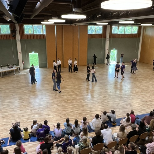 Tanzveranstaltung in einem gro&szlig;en Raum: Teilnehmer tanzen auf dem Holzfu&szlig;boden, Publikum sitzt und applaudiert.