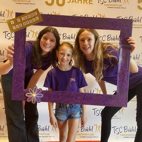 Drei Personen posieren in lila Shirts vor einer festlichen Hintergrundwand zum 50-j&auml;hrigen Jubil&auml;um des TSC Br&uuml;hl.