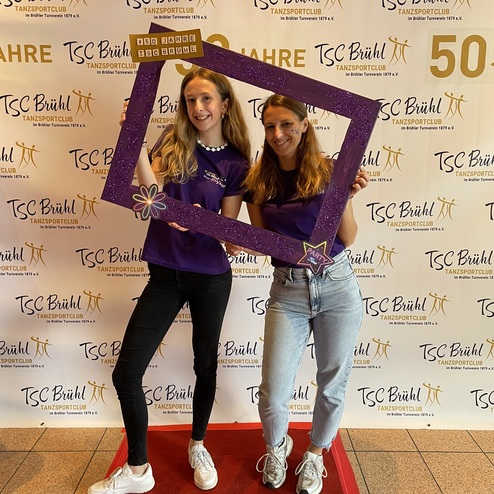 Zwei Frauen posieren mit einem lila Bilderrahmen vor einem Banner des TSC Br&uuml;hl, gekleidet in lila T-Shirts.