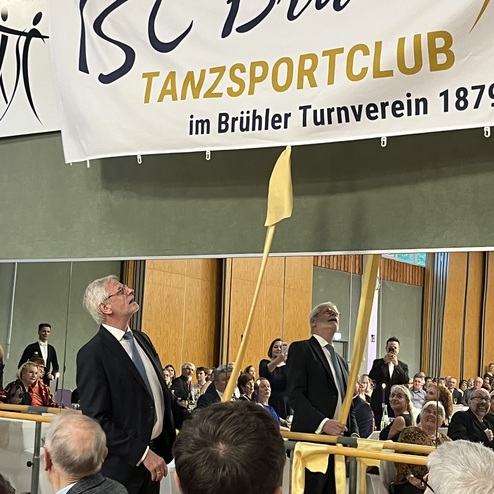 Zwei M&auml;nner in Anz&uuml;gen hissen ein Banner des TSC Br&uuml;hl w&auml;hrend einer Veranstaltung vor einem Publikum.