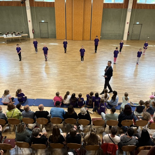 Eine Gruppe von T&auml;nzern in lila T-Shirts f&uuml;hrt eine Auff&uuml;hrung in einem gro&szlig;en Saal vor aufmerksamen Zuschauern auf.