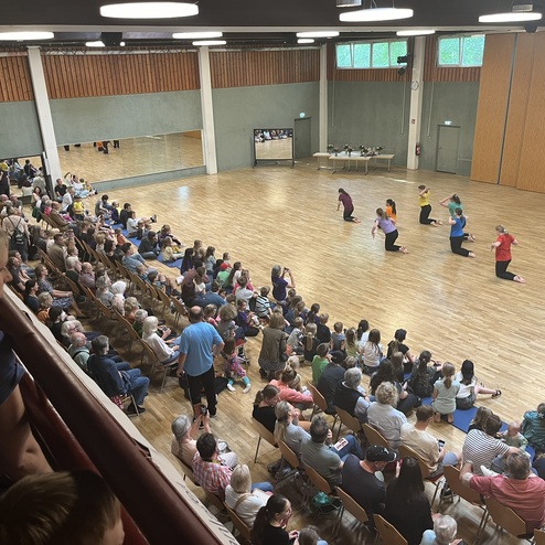 Tanzauff&uuml;hrung in einem gro&szlig;en Saal, umgeben von Zuschauern, mit T&auml;nzern in bunten Shirts auf dem Holzfu&szlig;boden.
