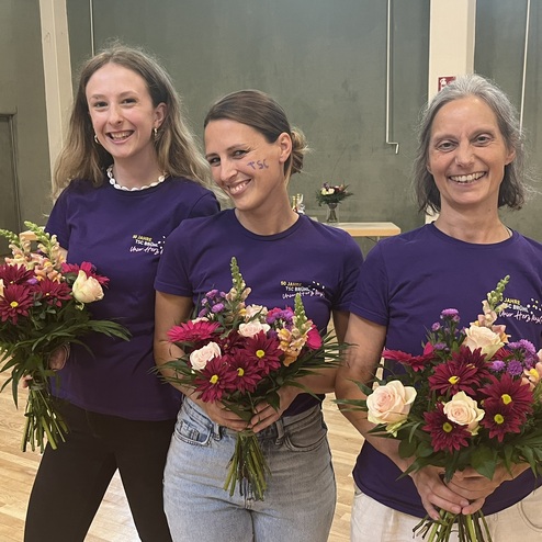 Drei Frauen in lila T-Shirts halten bunte Blumenstr&auml;u&szlig;e in einem gut beleuchteten Raum.