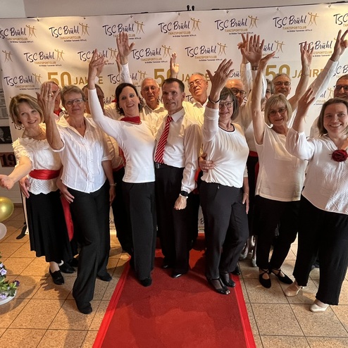 Gruppenselfie von T&auml;nzern in festlicher Kleidung, die vor einem Banner zum 50-j&auml;hrigen Jubil&auml;um posieren.
