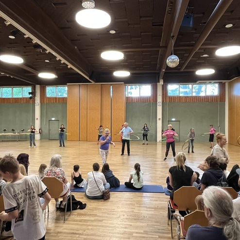 Gruppe von Menschen in einem Tanzsaal, die Hula-Hoop-Reifen in verschiedenen Farben nutzen, Publikum sitzt auf St&uuml;hlen.