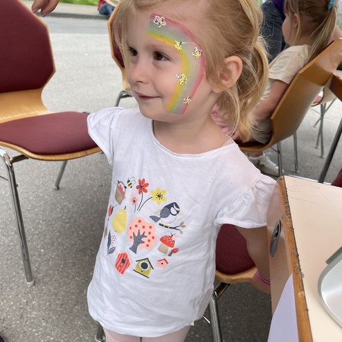 Kleines M&auml;dchen mit einem Regenbogen-Gesichtsmal in einem wei&szlig;en T-Shirt steht an einem Tisch, umgeben von St&uuml;hlen.