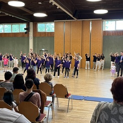 Tanzende Kinder und Jugendliche in lila T-Shirts zeigen eine einstudierte Choreografie in einem gro&szlig;en Saal.