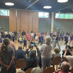 Viele Kinder und Erwachsene tanzen gemeinsam in einem Saal, w&auml;hrend Zuschauer auf St&uuml;hlen zuschauen und filmen.