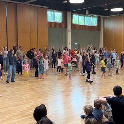 Versammlung von Kindern und Erwachsenen, die in einem hellen Raum gemeinsam tanzen und sich bewegen.