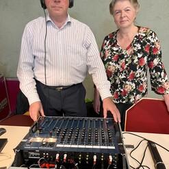 Zwei Personen stehen neben einem Audiomischpult, eins tr&auml;gt Kopfh&ouml;rer, beide blicken in die Kamera.