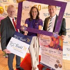 Drei Personen posieren vor einem Banner zur 50-Jahr-Feier des Tanzsportclubs Br&uuml;hl, halten Schilder und Plakate.